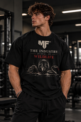 Male fitness model wearing a black oversized  MF graphic gym T-shirt featuring ‘The Industry Is Full of Wildlife’ slogan with wolf, rat, and snake artwork. MUSCLE FACTORY apparel brand. 